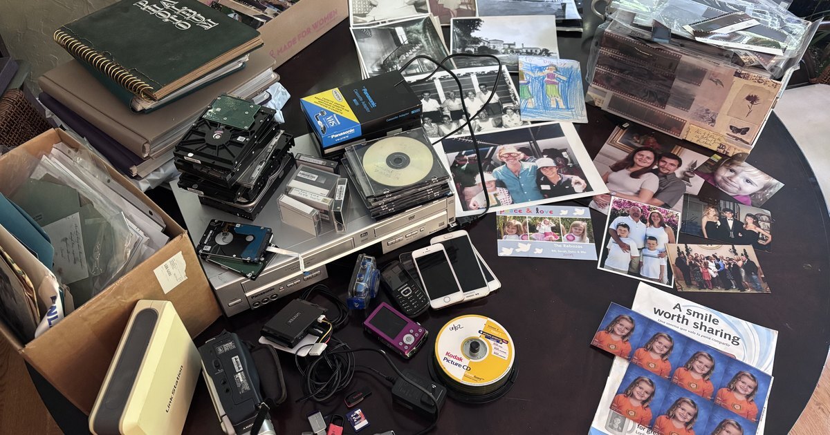  A dining table covered in one family's photos and media from every era: photo albums, a shoebox of prints, black-and-white photos, negatives, bare hard drives, CDs, camcorder tapes, a VHS and DVD player, a camcorder, old phones, and USB sticks.